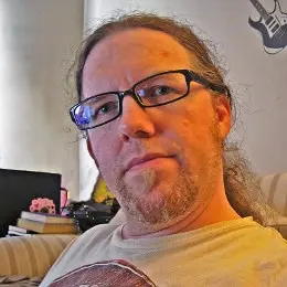 A man with long hair, glasses, and a short beard sits indoors on a striped couch. Behind him are a stack of books, a laptop, and a wall decorated with a guitar illustration.
