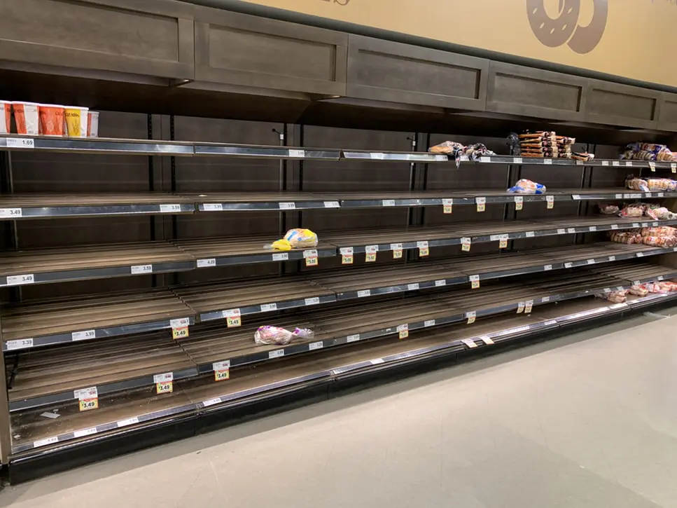 Nearly empty supermarket shelves with only a few remaining loaves of bread, buns, and packaged bakery items scattered across highlight the challenges of business survival during a pandemic, as most shelves are bare due to shortages or high demand.