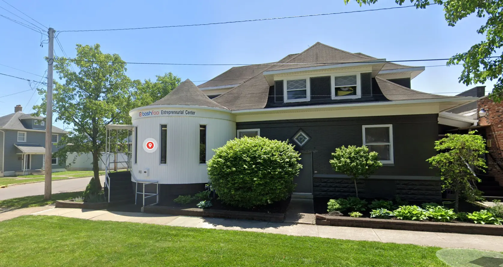 A two-story dark gray house with white trim, featuring a modern circular entrance labeled The Shop: Entrepreneurial Center, has been empowering small businesses for 25 years, surrounded by green shrubs and trees on a sunny day.