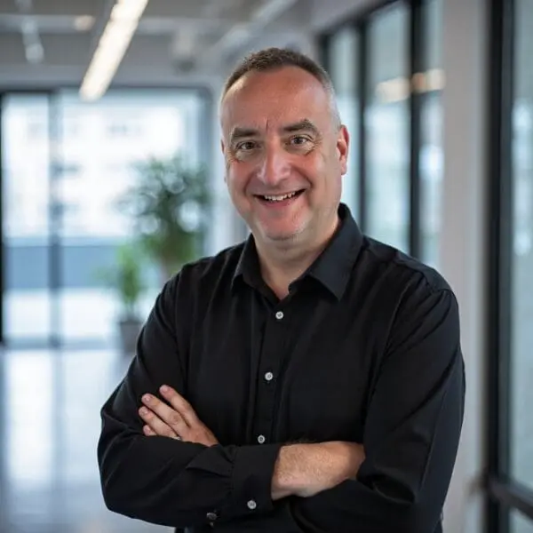 Mike McDermott, a man with short hair in a black button-up shirt, stands indoors with arms crossed, smiling at the camera. Large windows and a blurred office setting with plants fill the background.